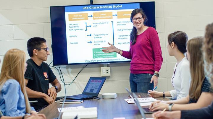 A female professor stands facing 5 students with laptops sitting at a table. She points at a screen.